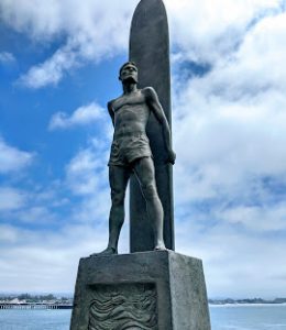 A large bronze statue of a muscular man in board shorts in front on a long board